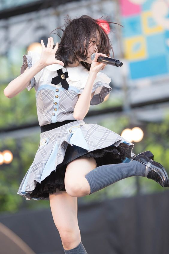 a young woman with short dark hair, wearing a white and blue plaid dress with a black collar and black lace trim, is captured midjump on a stage. she holds a microphone in her right hand and her left hand is raised. she is wearing kneehigh gray socks and black shoes. the background is blurred, suggesting a crowd and outdoor concert setting. the lighting is bright and natural, highlighting the performer's energetic movement.