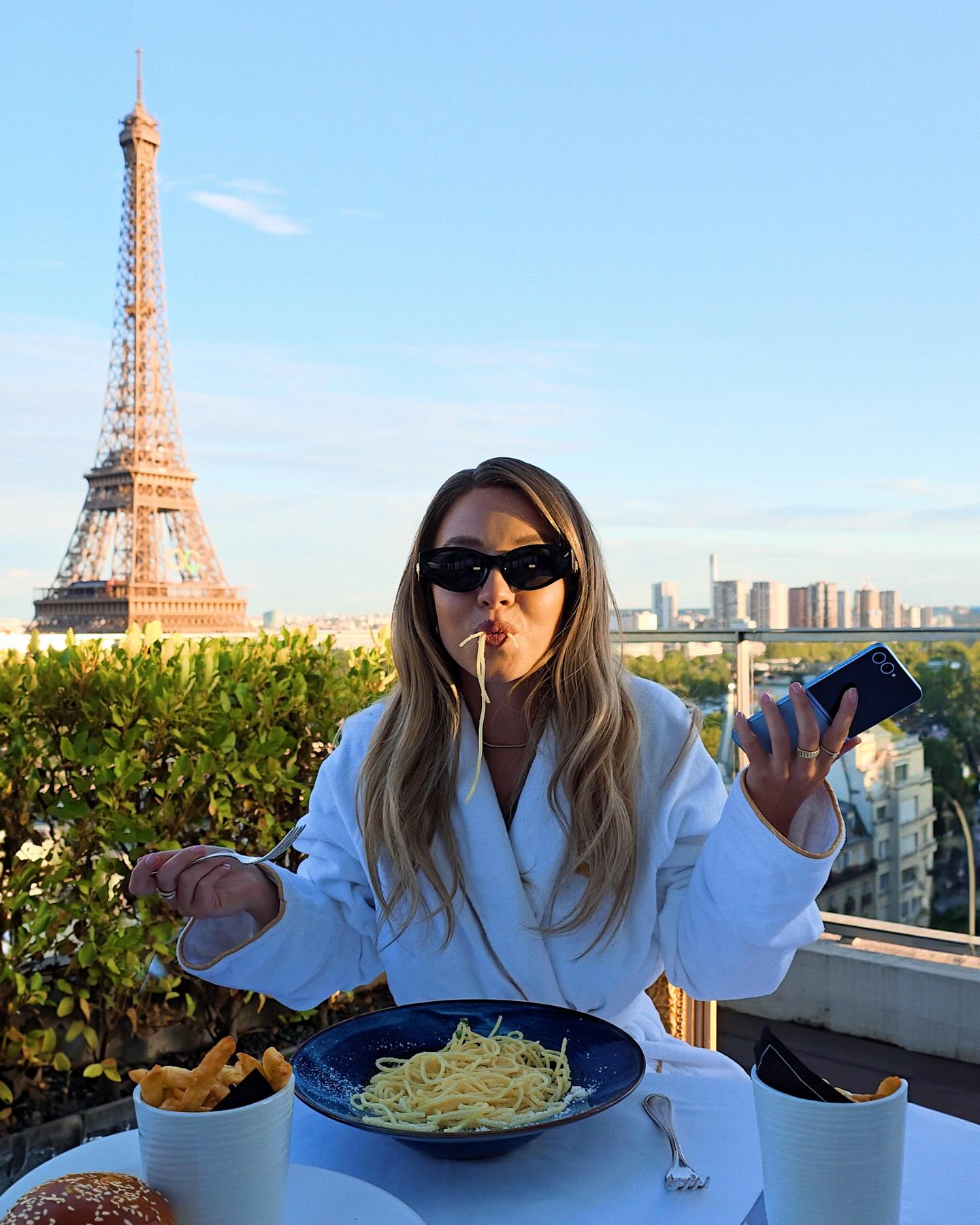 a blonde woman wearing sunglasses and a white robe is sitting on a rooftop restaurant in paris, enjoying a plate of spaghetti cacio e pepe.  the eiffel tower is visible in the background, showcasing a stunning cityscape. she is holding a phone in one hand and a fork in the other, and french fries are on the table. the scene exudes luxury and relaxation.