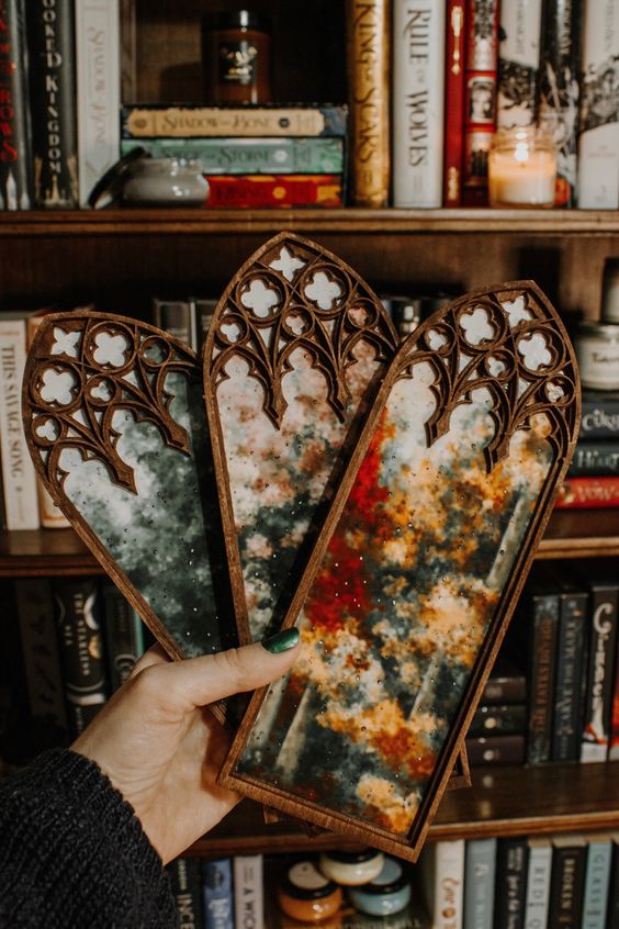 a hand holds three gothic stained glass windows, the windows are framed in wood and feature a cosmic design. the windows are in front of a bookshelf filled with books.