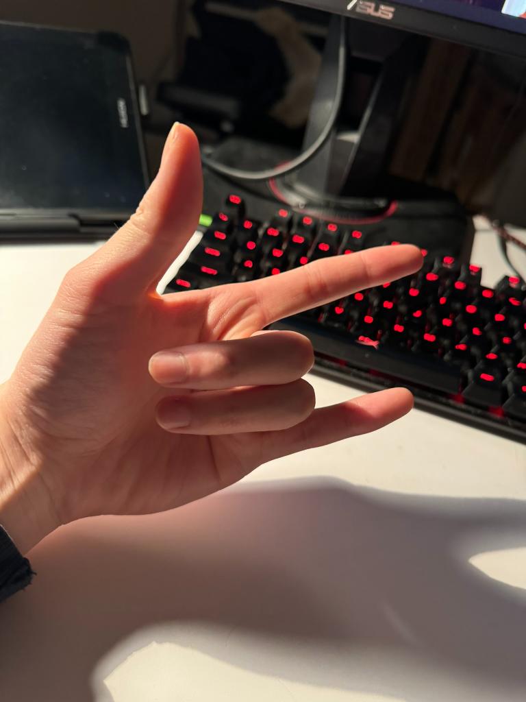 a left hand making the rock and roll gesture is seen over a white desk with a black keyboard with red lighting and a black monitor on the background. the hand is illuminated by sunlight.