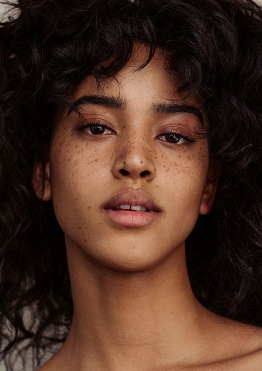 a closeup portrait of a beautiful woman with freckles, dark curly hair, and dark eyes. she is looking directly at the camera. her skin is smooth and her lips are slightly parted. the image focuses on her face and upper body, with the background blurred out.