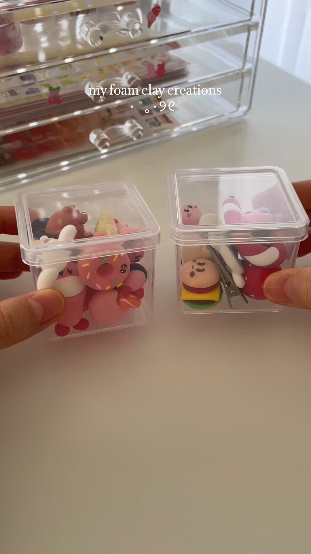the image shows two small, clear plastic boxes filled with foam clay creations. the left box contains multiple pieces, including pink donuts and cartoon figures. the right box holds a burger, another cartoon figure, and a silver hairpin. both boxes are being held by hands. behind the boxes, there is an acrylic organizer filled with decorative items and a text overlay saying "my foam clay creations  9€".