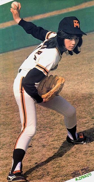 a color image of a young woman in a baseball uniform winding up to pitch a baseball. she is wearing a white uniform with black and orange trim, a black baseball cap with a logo, white socks, and black and white cleats. she has long black hair and is looking intently at her target. the background is a green baseball field. the image appears to be from the 1970s or 1980s.