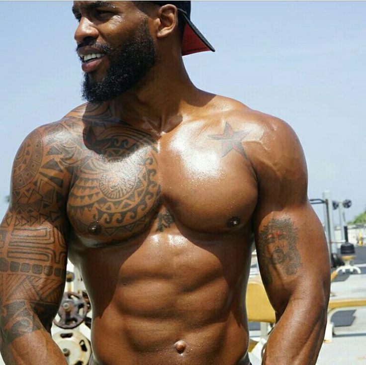 a shirtless man with tattoos on his chest and arm is flexing his muscles in an outdoor gym. he is looking at the camera with a confident expression. he has a welldefined six pack, and his biceps and triceps are bulging. he has a black beard and a black baseball cap.