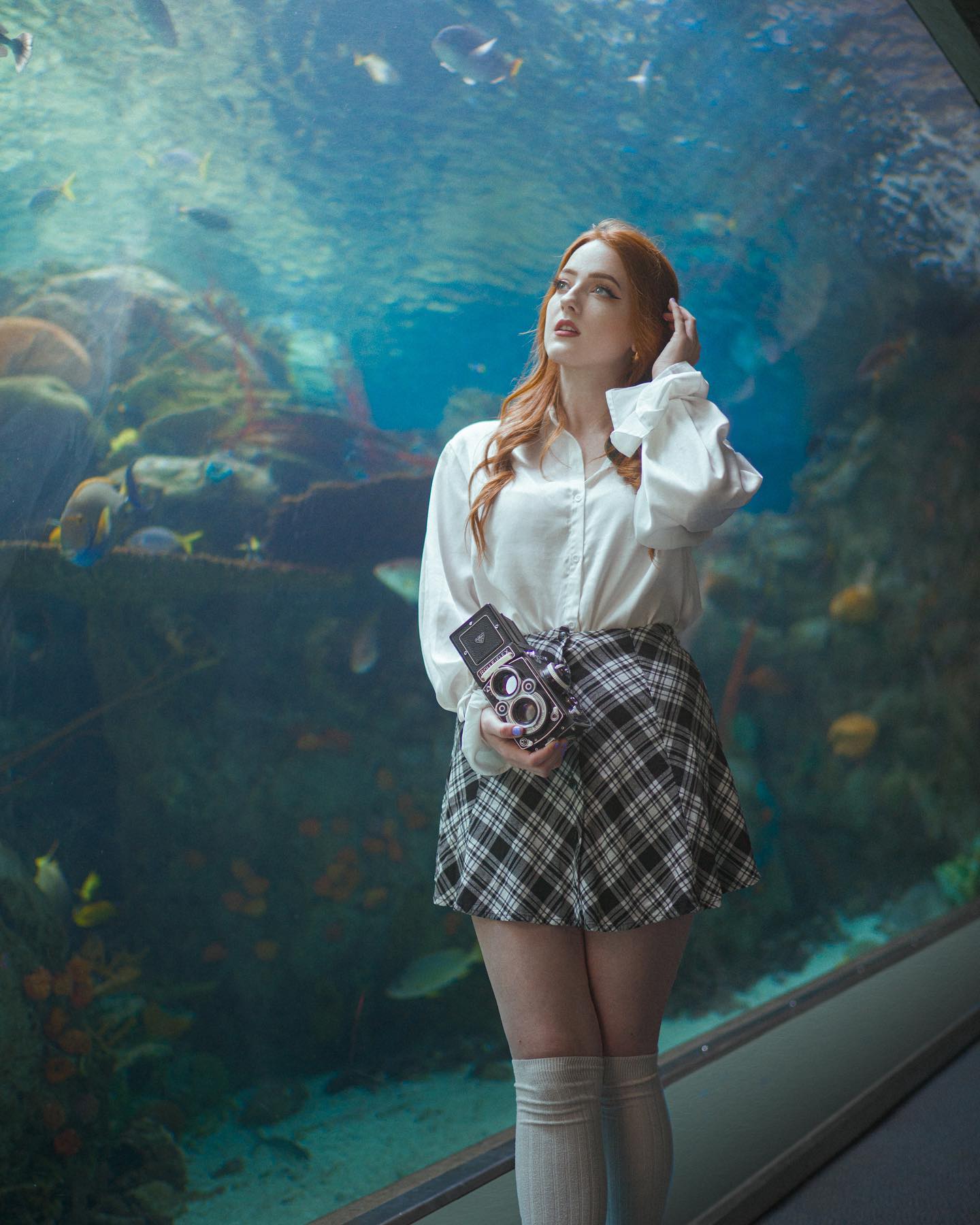 a young woman with long red hair stands in front of a large aquarium tank. she is wearing a white blouse, black and white checkered skirt, and white kneehigh socks. she is holding a vintage camera in her hands and looking up at the fish swimming in the tank. the lighting is soft and diffused, creating a dreamy and ethereal atmosphere.