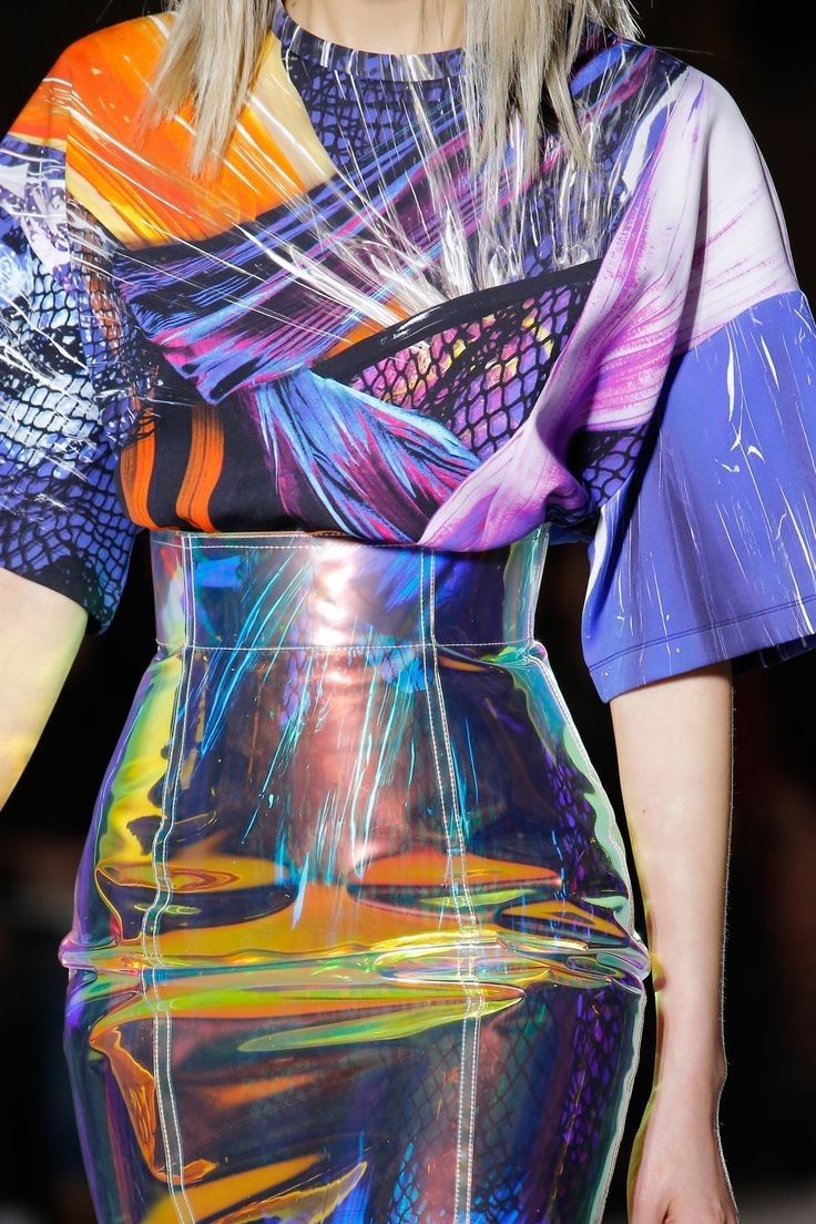 a model is wearing a colorful holographic dress on a runway. the top of the dress is patterned with orange, purple, blue, and black designs, while the bottom is a reflective holographic material with hues of blue, green, orange, and purple. the dress has short sleeves and a belt that accentuates the waist.