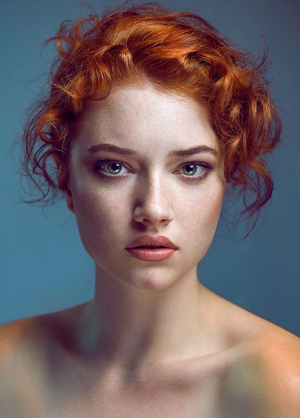 a studio portrait of a young woman with bright red, styled curly hair and freckles. she is facing forward, looking directly at the camera with a neutral expression. she has fair skin, and her shoulders are bare. the background is a muted light blue color. the lighting is soft, accentuating her features and creating a natural, yet polished look.