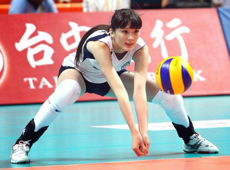 a female volleyball player in a white jersey and black shorts is diving to spike the ball. she is wearing knee pads and white socks with black shoes. she is playing on an indoor court, and the ball is in midair. the background is a red wall with chinese characters. the focus is on the player and the ball. the image captures the intensity of the game.