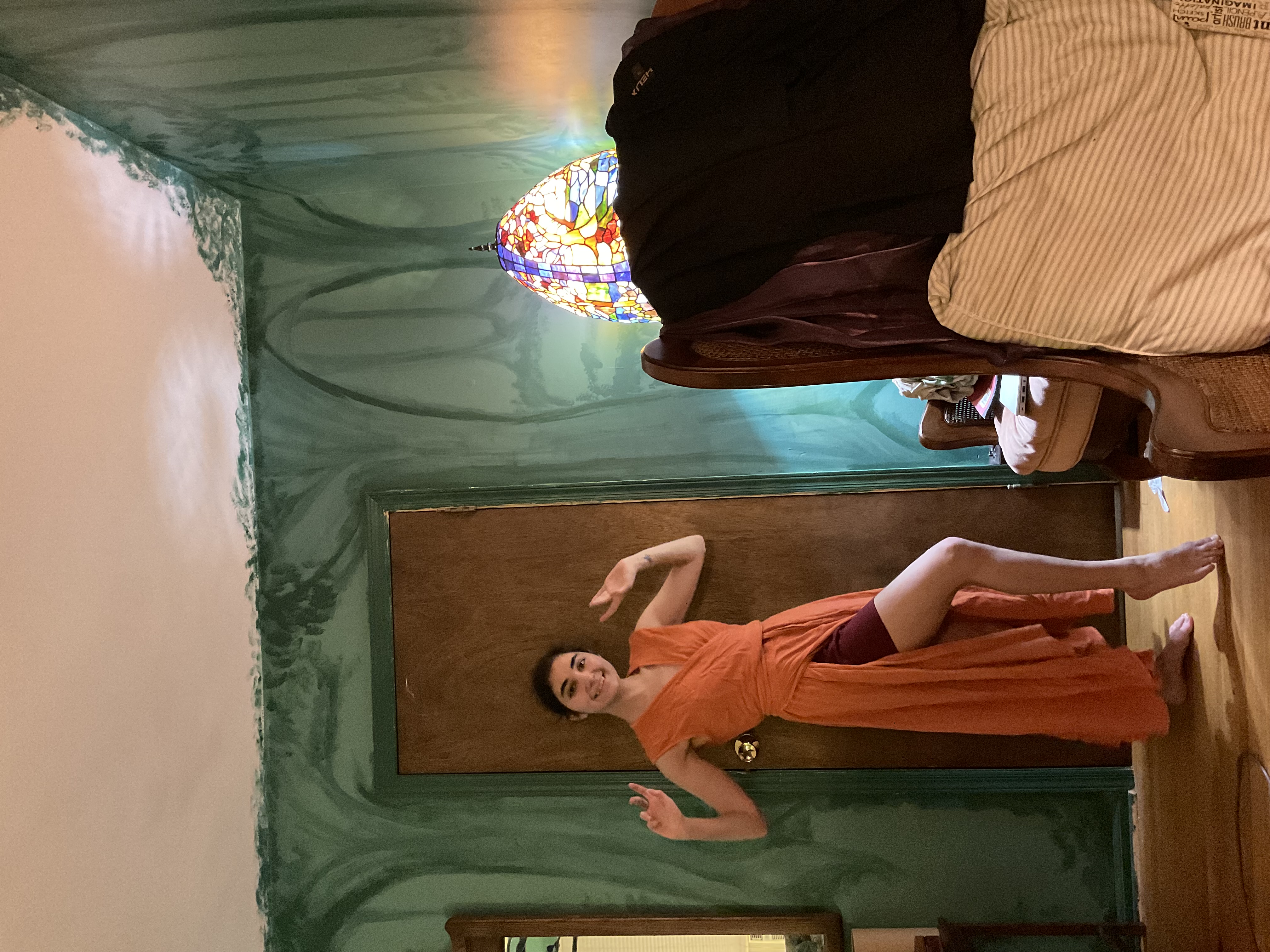 a young woman in a flowing orange wrap dress is posing and seemingly dancing in a room with a striking green wall mural depicting a forest scene.  the dress is highlow, revealing dark leggings or shorts underneath. she's barefoot.  a vintagestyle stainedglass lamp sits on a wicker armchair in the background. the room has a slightly bohemian and artistic feel. the overall mood is relaxed and expressive.