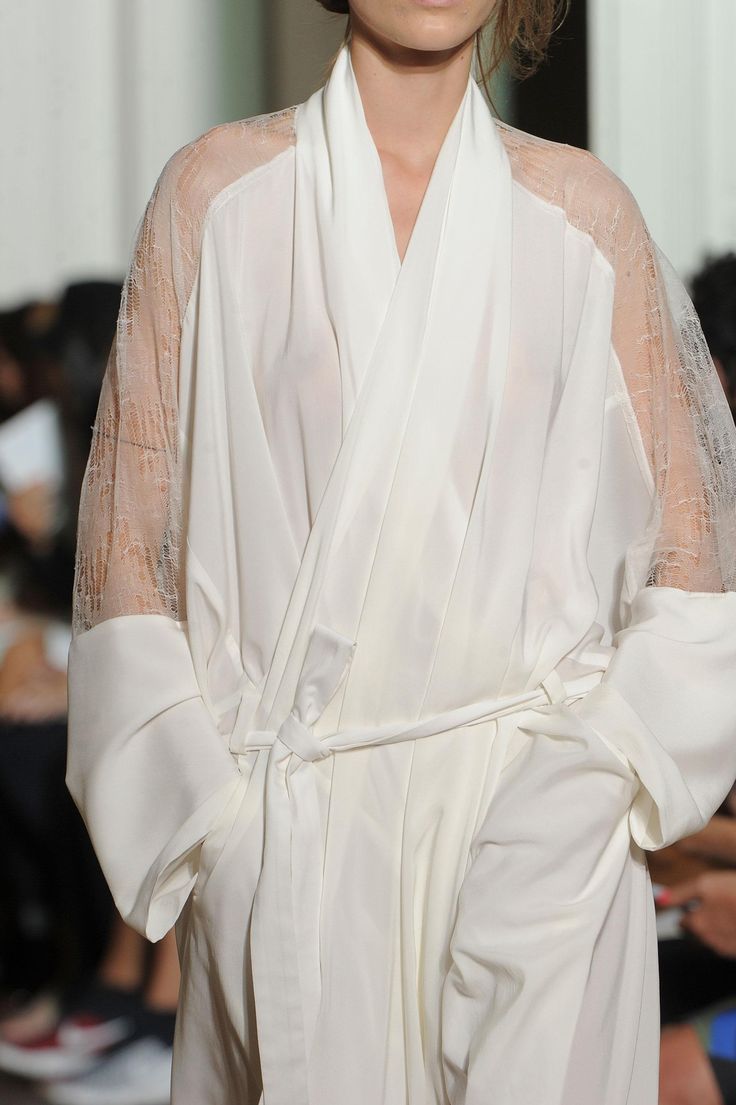 a model in a white gown with lace sleeves walks the runway. the gown is cinched at the waist with a simple belt. the model is looking down and slightly to the left.