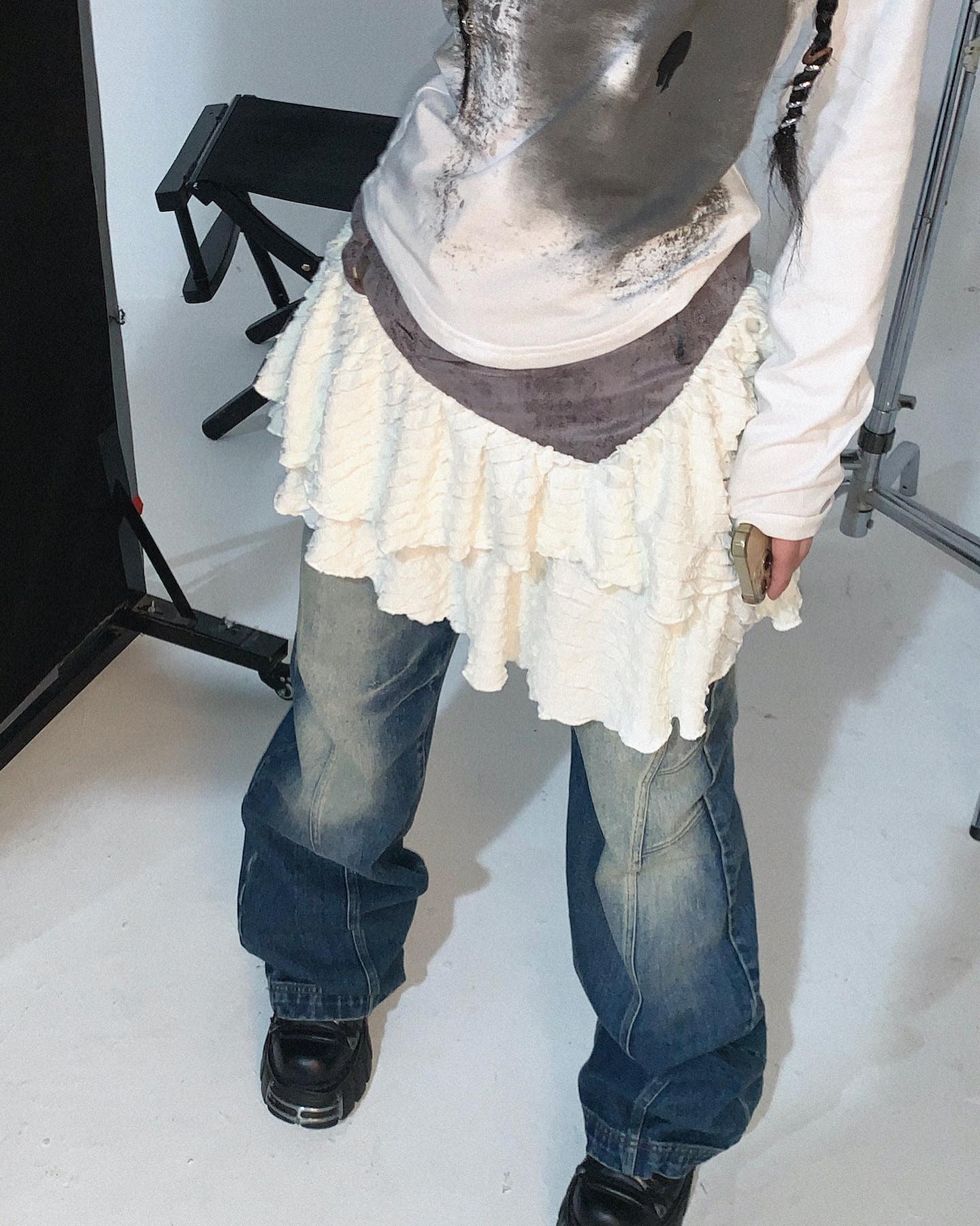 a closeup shot of a fashion model showcasing a layered ruffled white skirt over denim jeans. the model is wearing an oversized white longsleeved shirt with a distressed look and a dark gray waistband under the skirt. the outfit is styled in a grunge and streetwear aesthetic. the model's footwear includes platform boots. the background is minimalistic with a white backdrop and a folding chair. the focus is on the outfit's details and textures.