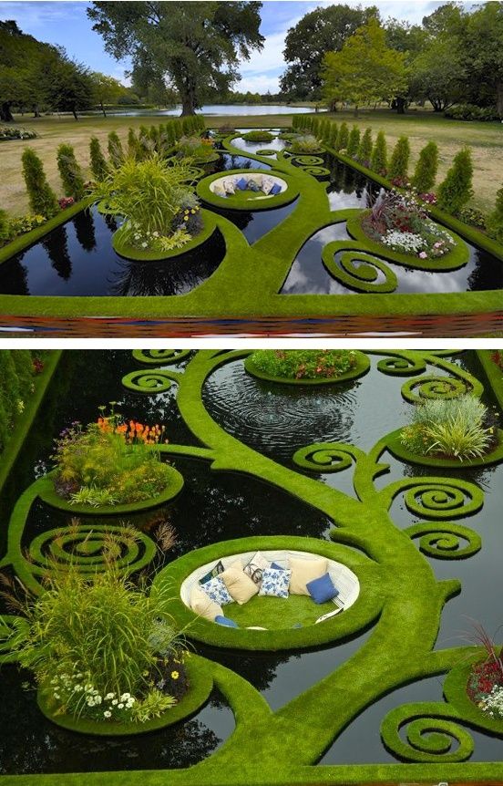 the image showcases a beautifully designed garden with a unique water feature. the pond is surrounded by lush green grass that forms a swirling, treelike pattern, creating a visual masterpiece. small islands of grass are adorned with various ornamental plants and flowers, adding pops of color and interest. a circular seating area with comfortable pillows offers a relaxing space within the garden. the topiary and wellmaintained hedges contribute to the overall manicured and decorative aesthetic of the outdoor space. in the background, mature trees and a lake can be seen, enhancing the natural beauty of the garden.