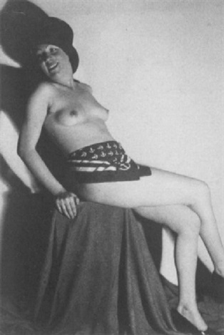 a black and white photograph shows a nude woman seated on a draped fabric. she wears a top hat and a silk scarf around her waist. her expression is slightly mischievous or amused. the background is plain. the image has a vintage feel, suggesting it might date from the 1920s or 1930s. the lighting and composition are classic elements of studio portraiture.