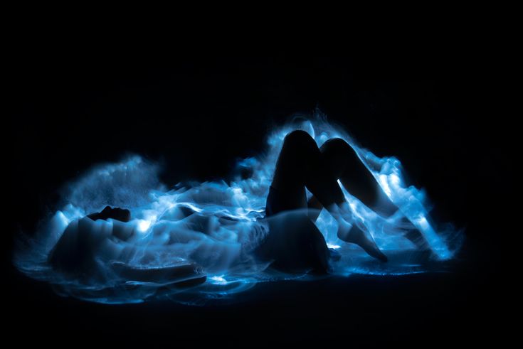 the image shows the silhouette of a woman lying on her side in a body of water that is glowing with bioluminescence. the water emits a bright, ethereal blue light, creating a surreal and dreamlike atmosphere.  the woman's form is mostly dark, contrasting strongly with the vibrant blue glow surrounding her. the overall effect is one of mystery and beauty, suggesting a magical or fantastical scene. the background is completely black, focusing all attention on the luminous water and the figure within it.