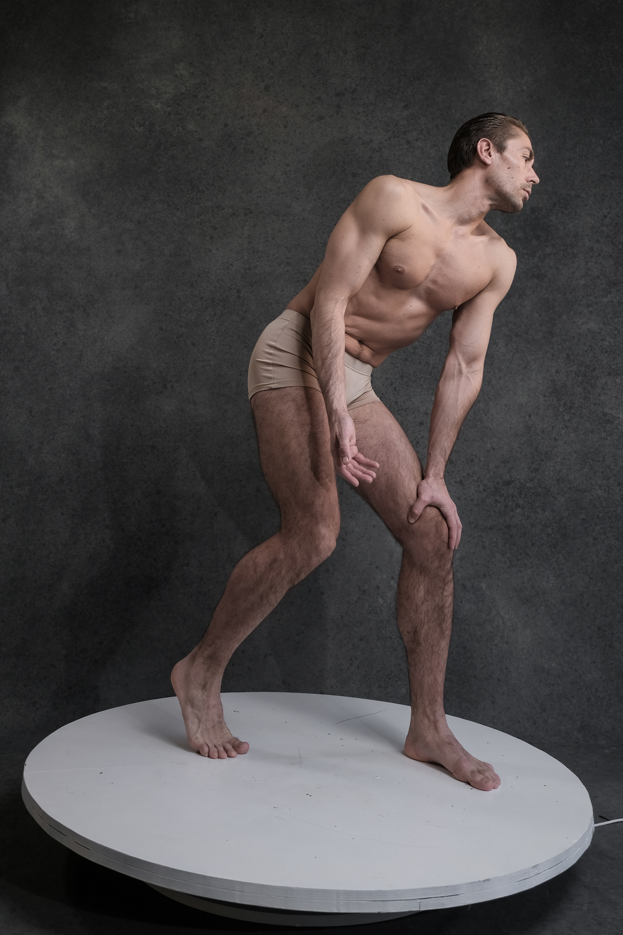the image showcases a muscular man in beige underwear, striking a dynamic pose on a white rotating platform against a textured gray backdrop.  he stands with one leg extended and the other bent, torso leaning forward, and hands reaching down. the soft lighting illuminates his physique and creates a sense of depth.