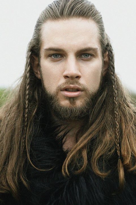 a portrait of a man with long blonde hair and a beard, styled with braids on the sides. he has blue eyes and is wearing fur clothing, giving him a vikinglike appearance.