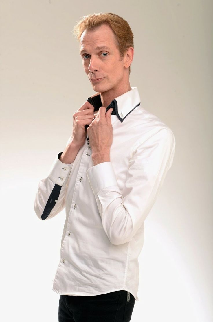 a portrait of a man wearing a white shirt with a black collar. the man is looking at the camera, smiling, and has blonde hair. the background is white. he is standing with his arms crossed.