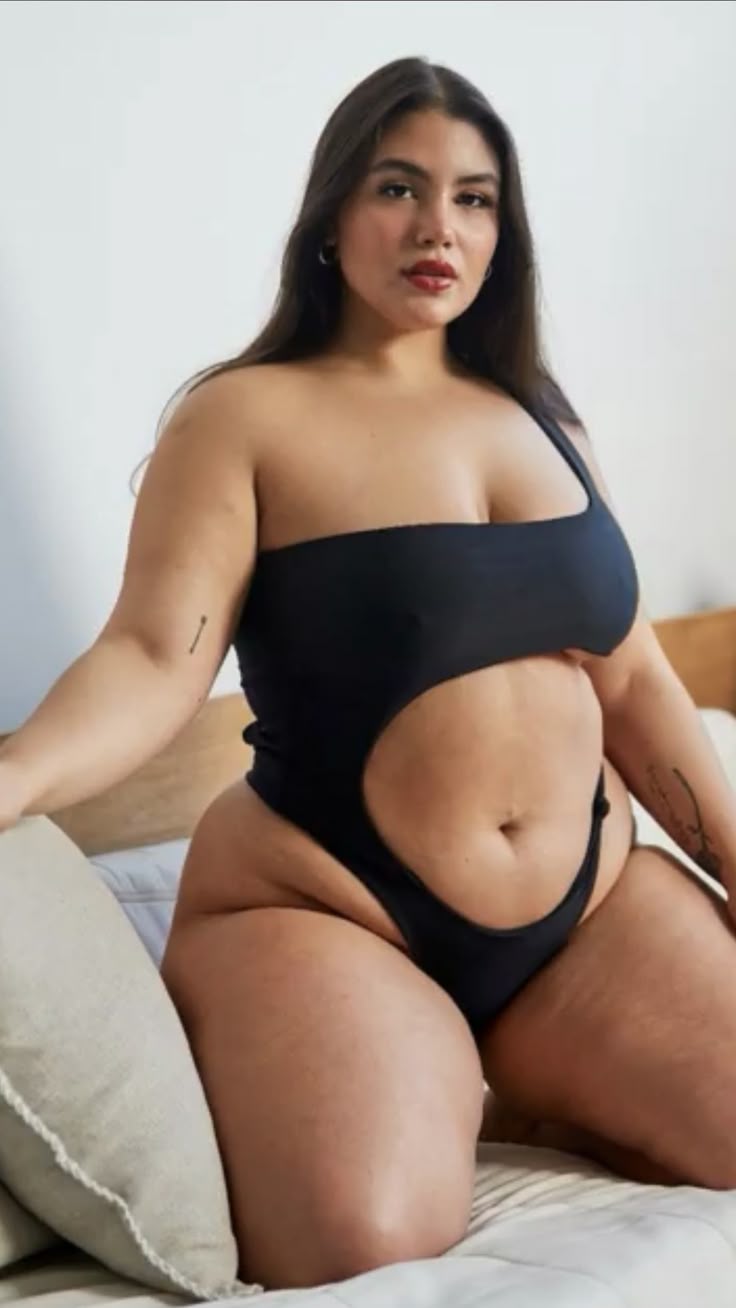 a plussize woman with curves is sitting on a bed in a black onepiece swimsuit. she appears confident and comfortable. the woman has dark hair and is wearing red lipstick. the background includes a pillow and bedding. she has tattoos on her arms.