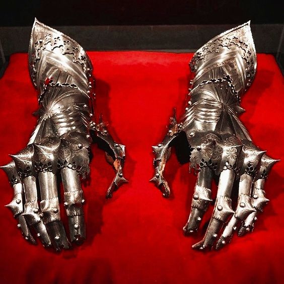 the image shows a pair of intricately crafted metal gauntlets from a suit of armor, displayed on a red surface, likely within a museum. the gauntlets are made of steel with a dull, metallic finish. they are designed to protect the wearer's hands and forearms in battle, with articulated plates for flexibility and sharp spikes on the knuckles for offensive purposes. the red background provides a stark contrast and emphasizes the metallic details of the gauntlets.