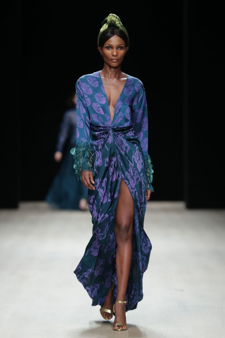a darkskinned model walks the runway wearing a teal and purple floral print gown. the gown is longsleeved, has a vneck and a high leg slit. the model is also wearing a green headwrap and gold high heels. the background is dark and blurred. the overall style is sophisticated and elegant.