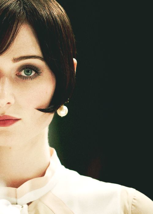 a woman with short black bobbed hair, bangs, red lipstick, pearl earrings and a white collared blouse looks at the camera. the background is dark and blurry.