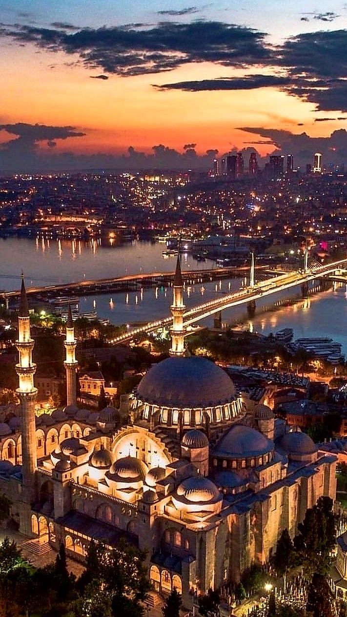 this image captures the stunning cityscape of istanbul, turkey, at dusk. the suleymaniye mosque is prominently featured, its illuminated domes and minarets standing out against the fading light. the architecture is classic ottoman style, and the scene includes a bridge spanning the waterway. the overall image showcases the urban beauty of istanbul during the golden hour, with the city lights beginning to twinkle as night approaches. the sunset colors are reflected on the water, enhancing the picturesque view.