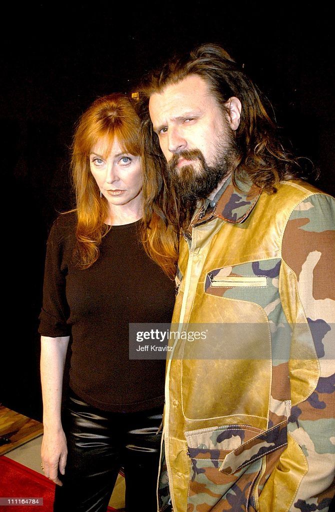 a man and woman stand together with their arms around each other in front of a dark background.  the man is wearing a camouflage jacket and the woman is wearing a black shirt and pants. they both have long brown hair.  the man is looking at the camera and the woman is looking off to the side.