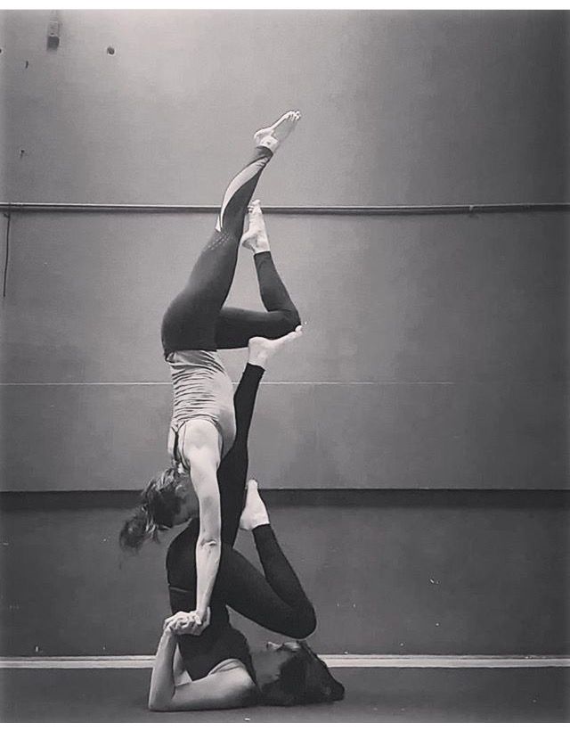 two women are performing an acrobatic yoga pose in a studio. one woman is standing on her hands, with her legs up in the air, while the other woman is balancing on her feet, holding the first woman's legs. they are both smiling and looking at the camera. the image is in black and white.