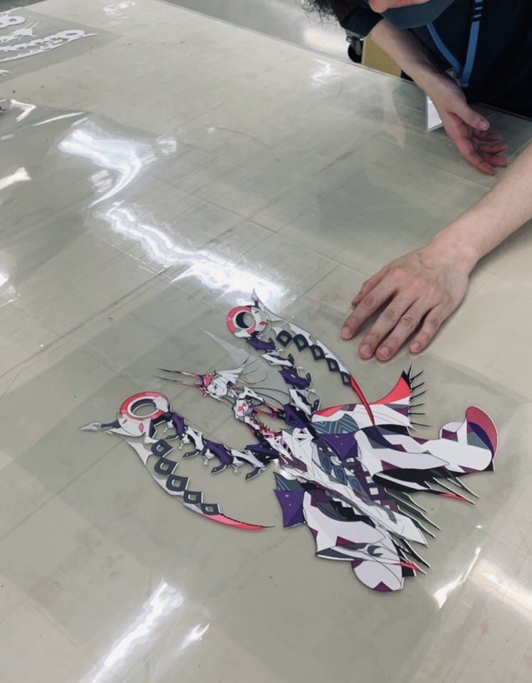 a topdown perspective of an artist working on a largescale print of a stylized, dynamic character. the print is laid out on a transparent sheet over a light table, revealing intricate details and vibrant colors in the design.  the artist's hands are carefully positioned on either side of the print, suggesting meticulous attention to its assembly.  the surrounding studio environment is clean and bright, further emphasizing the focus on the artwork.