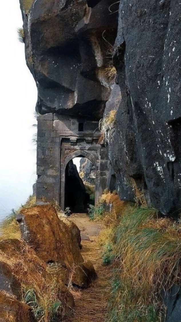 a stone doorway is situated between a rocky cliff face and a mossy rock formation. a path runs through the doorway. the surrounding landscape features grass and moss.