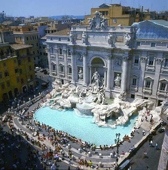 the image is a highangle, wide shot of the trevi fountain in rome, italy. the fountain is the central focus, displaying its intricate baroque architecture, numerous sculptures, and the turquoise water cascading down its tiered structure.  the surrounding piazza is populated with numerous tourists, giving a sense of scale and the popularity of this iconic landmark. the buildings behind the fountain showcase the urban architecture of rome. the scene is bathed in bright sunlight, indicative of a clear, summer day. the overall impression is one of bustling activity in a historic and beautiful setting.