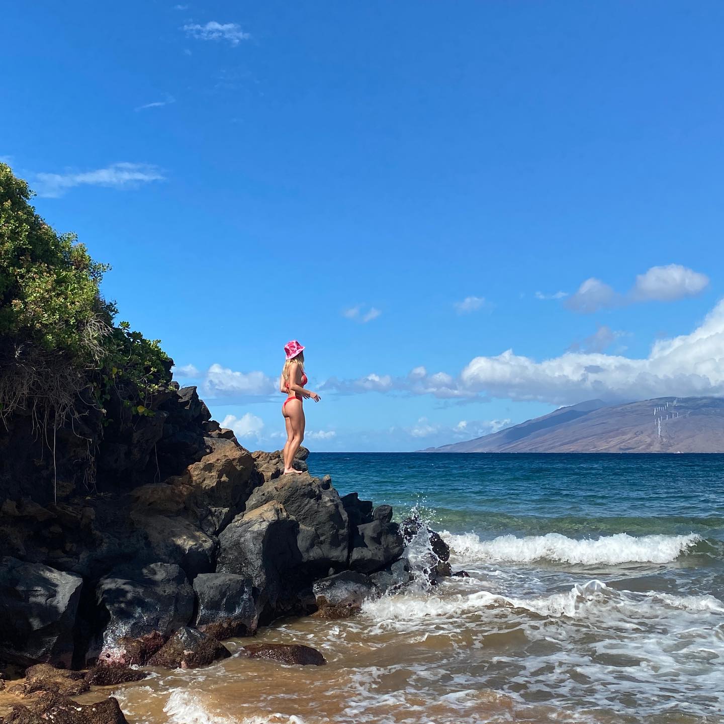 a woman in a red bikini and pink hat stands on a rocky outcrop overlooking a beautiful ocean scene. the ocean is a vibrant blue, with gentle waves lapping against the rocks. the sky is a clear, bright blue with fluffy white clouds. in the distance, a mountainous landscape can be seen. the overall impression is one of tropical paradise and tranquility.