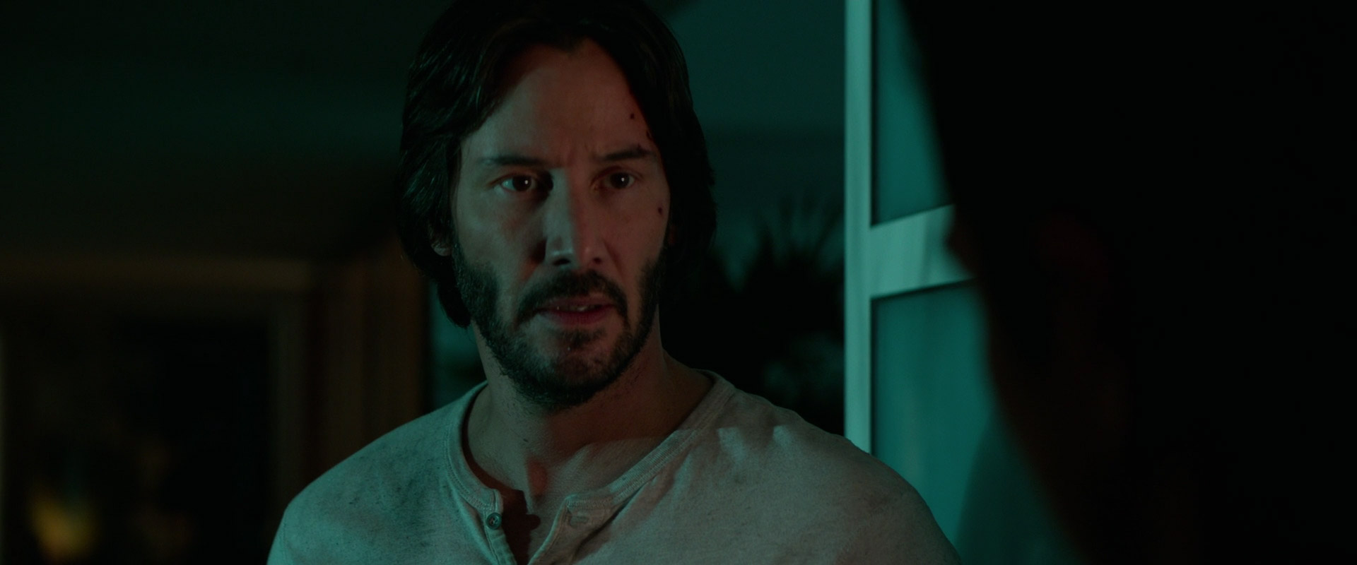 a closeup portrait of keanu reeves looking intensely into the camera. he's wearing a white tshirt and has a beard. the background is dark, and there's a window to his right.