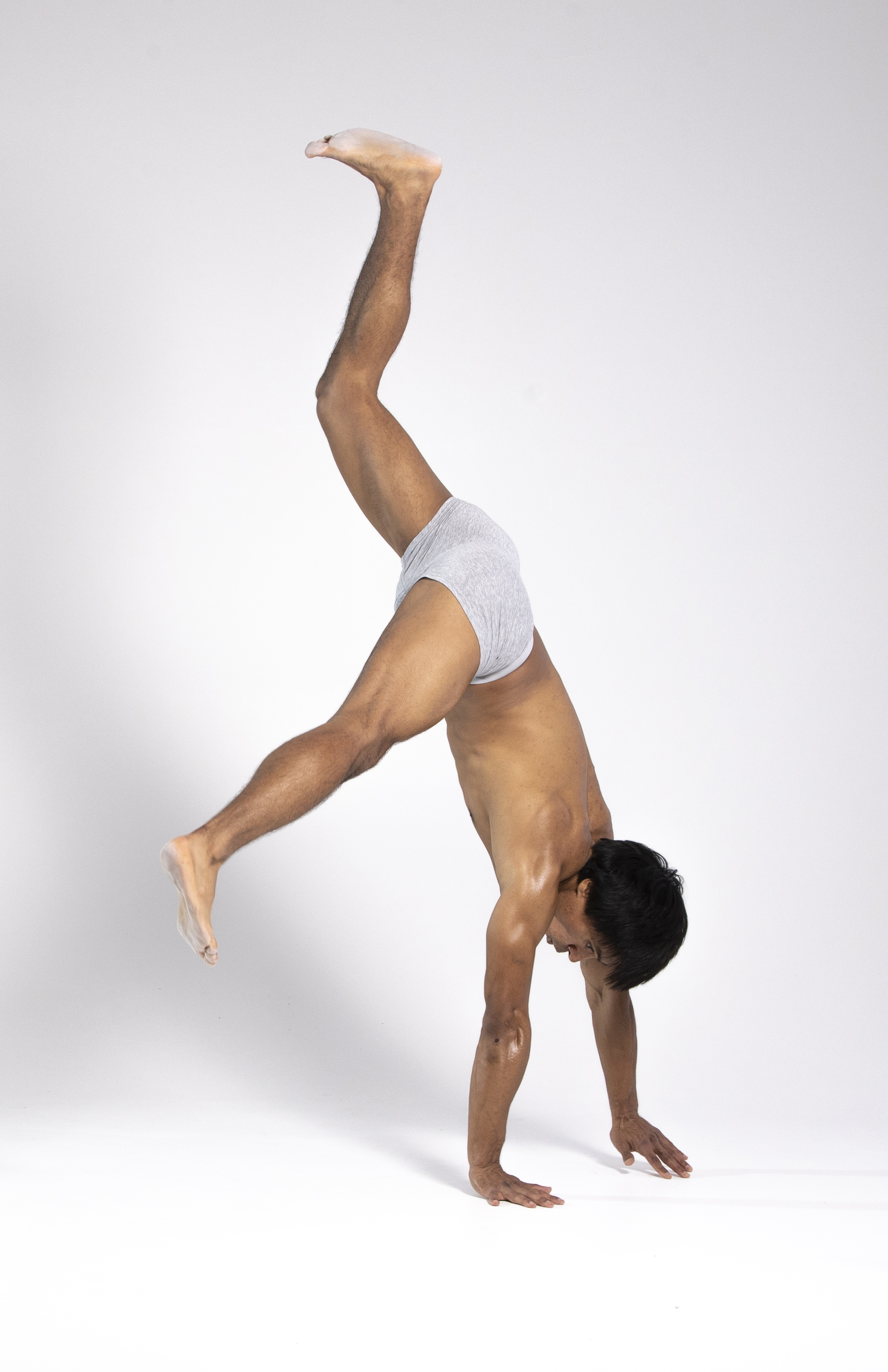 a man is performing a handstand in a studio setting, wearing underwear. he is showcasing his strength, flexibility, and bodyweight control. his body is inverted with one leg extended upward, while the other leg is bent at the knee. he is supporting his weight on his hands.