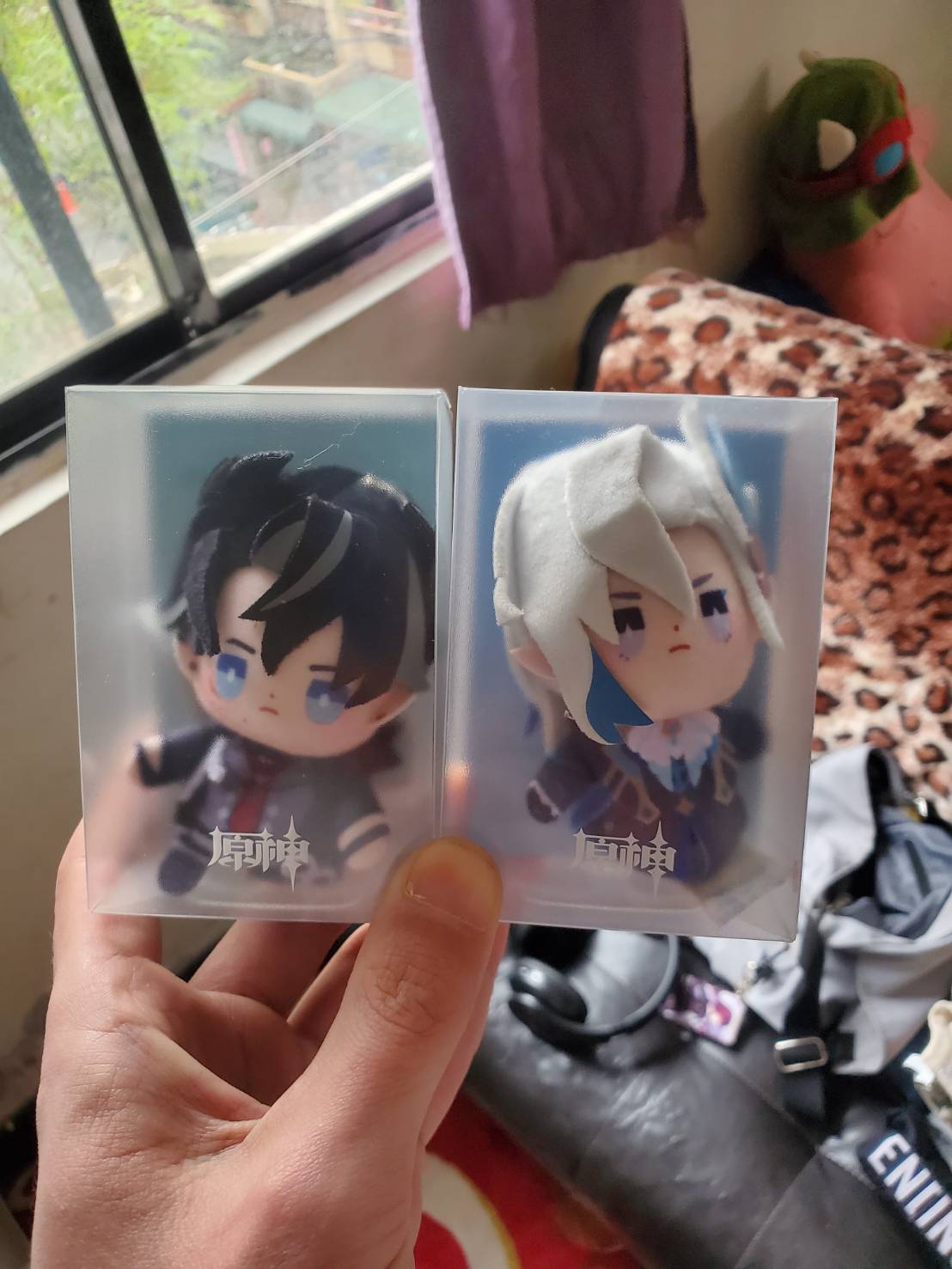 a hand holding two clear plastic packages containing anime figurines. the left package contains a blackhaired male character with a black and red outfit, and the right package contains a whitehaired female character with a white and blue outfit. both characters have large eyes and are smiling. the packages are labelled with the characters' names in japanese.