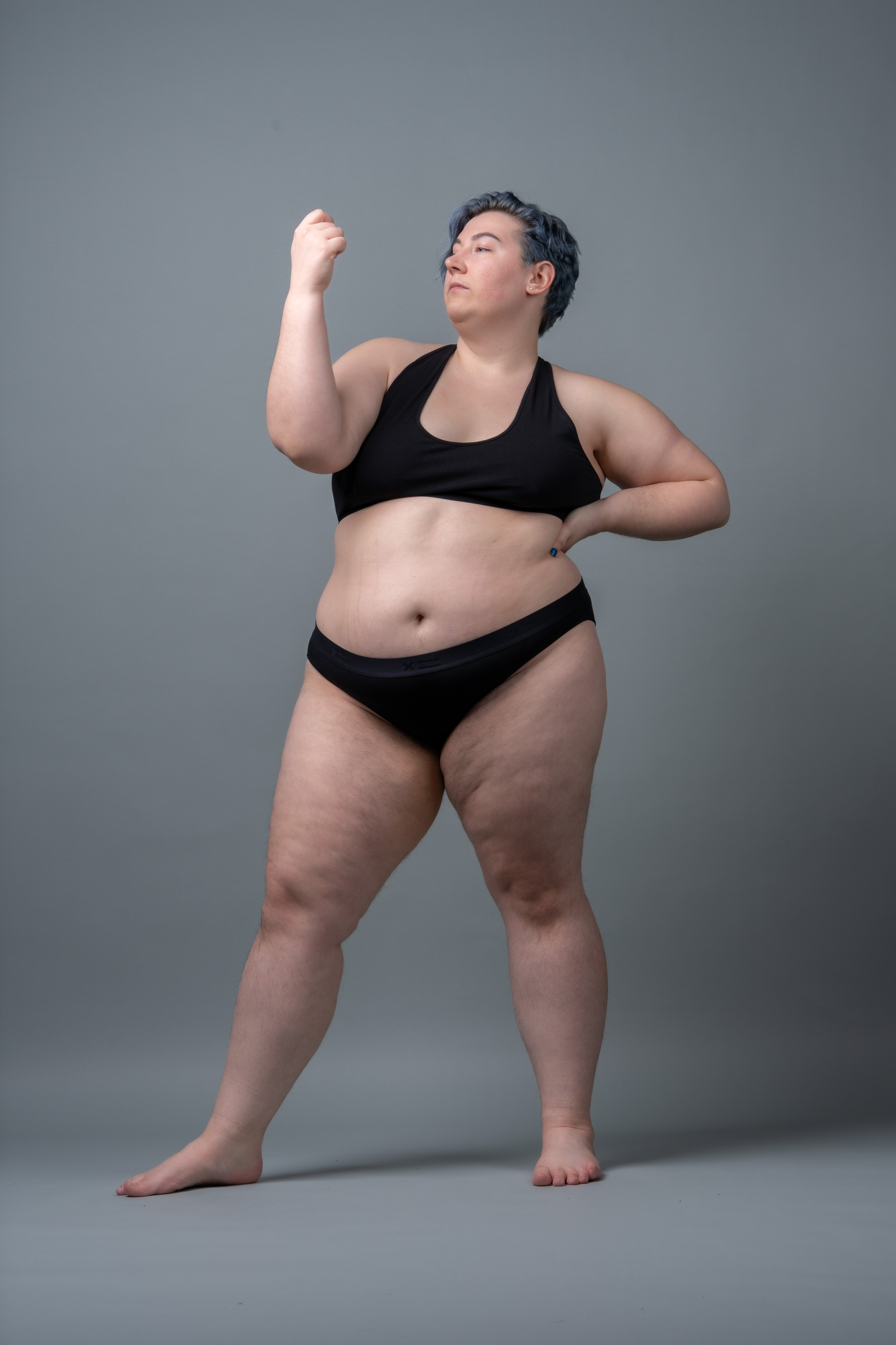 a plus size woman with blue hair stands confidently in a studio wearing black lingerie. she is barefoot and strikes a pose, conveying body positivity and selfassurance.
