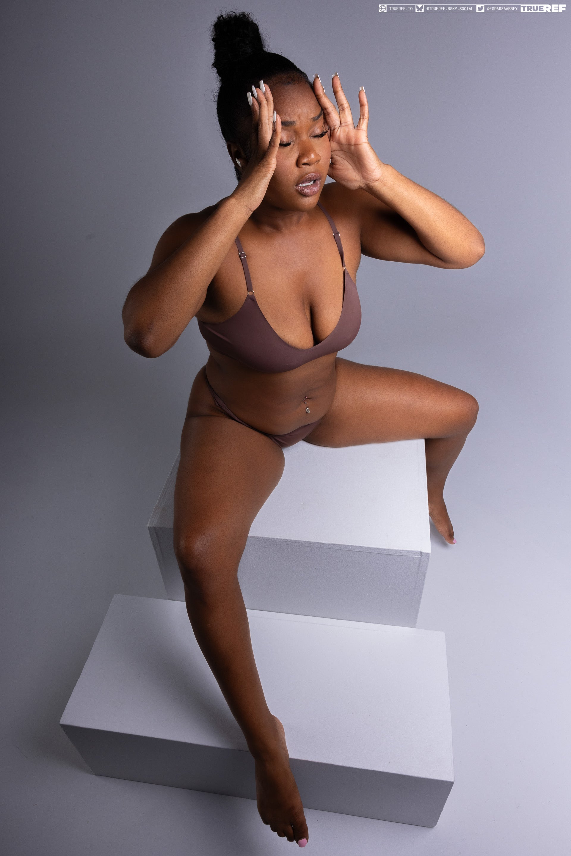 a darkskinned woman wearing brown lingerie sits atop a stacked white cube structure indoors, her expression conveying stress or pain as she holds her hands to her head.