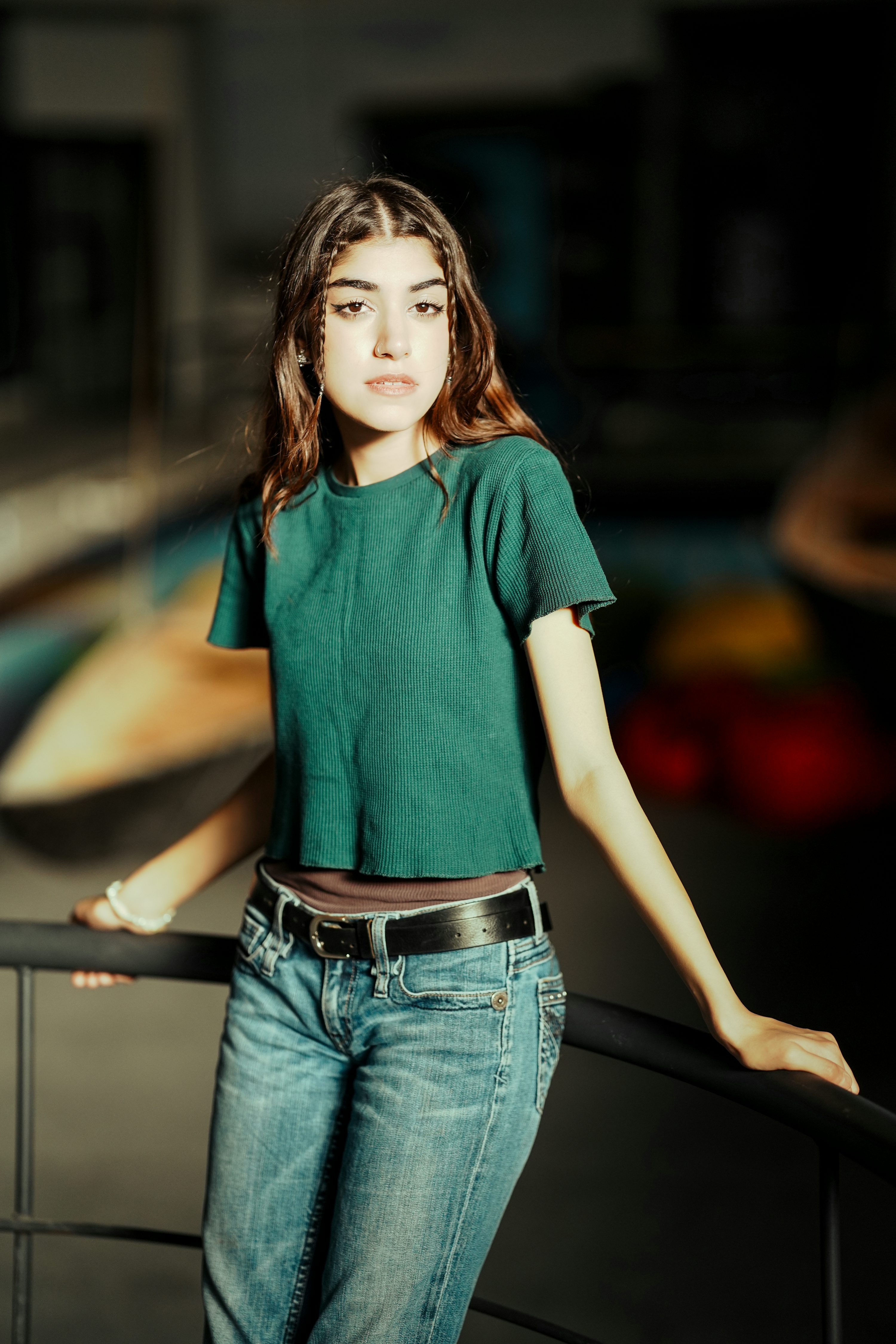 a young woman with braids in her hair stands indoors, leaning against a black metal railing. she is wearing a green shortsleeved top, blue jeans with a black belt, and a brown undershirt is visible. she has a serious expression on her face. the background is blurred.
