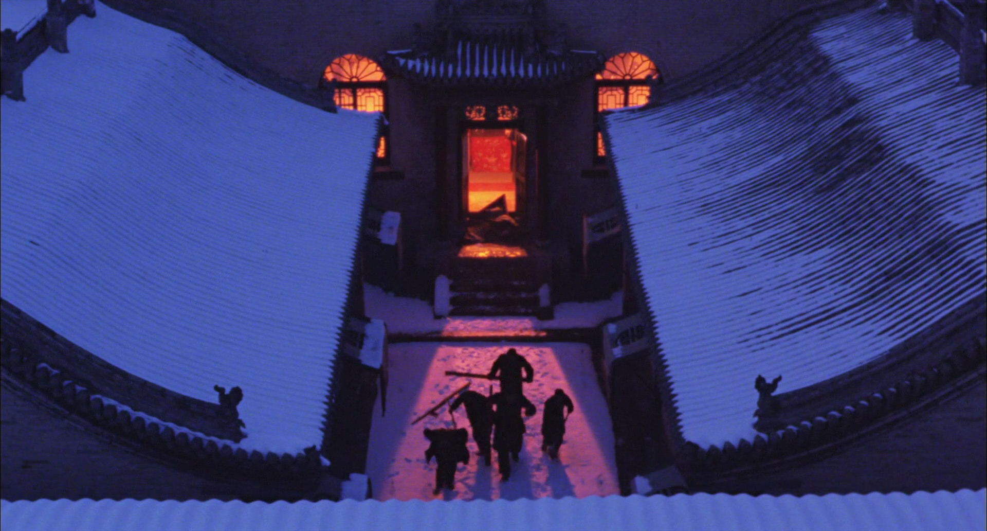 an aerial perspective of four people walking single file across a snowy courtyard at night.  the courtyard is surrounded by buildings, with the main building straight ahead.  the buildings have dark blue tiled roofs covered with a layer of snow. the main building is glowing warm orange, lit from within and by string lights.  footprints in the snow show the path the people took from the main entrance.