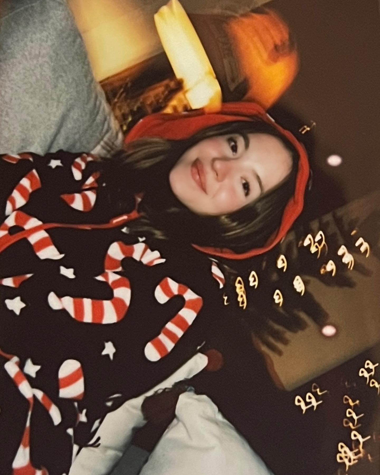 a young woman is lying down, wearing a black hoodie with a candy cane pattern and a red hood. she is smiling and appears happy. the image has a festive feel, likely due to the christmasthemed pajamas and blurred lights in the background, suggesting a holiday setting.