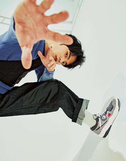 a young asian man in stylish clothing is captured midair, reaching towards the camera with an outstretched hand. he's wearing a blue sweater, dark grey cargo pants, white socks, and grey sneakers with white and red details. the low angle shot emphasizes the dynamism of the pose against a clean, white background.