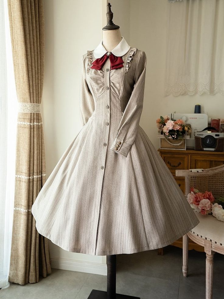 a longsleeved, vintagestyle dress in a beige and white pinstripe pattern is displayed on a mannequin in a welllit room. the dress features a full skirt, a high collar, and a row of buttons down the front. a red bow adds a touch of color to the neckline. the mannequin is positioned in front of a window with sheer curtains, and a wooden chair and table with flowers can be seen in the background.