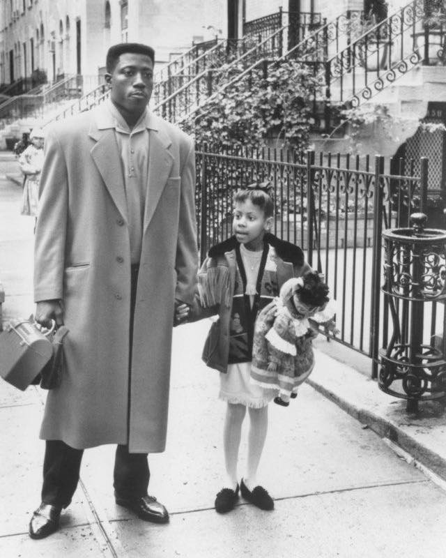 a black and white photograph shows a man and a young girl walking down a city street. the man is wearing a long overcoat and carrying a briefcase. the girl is wearing a dress and jacket. they are holding hands. the background is a typical urban setting with brownstone buildings and a wroughtiron fence.  the overall style suggests a film still or a photograph from the 1990s.