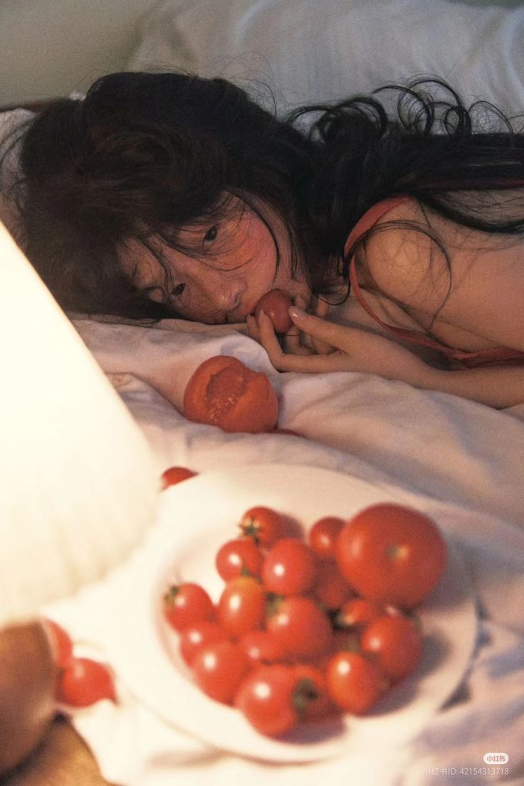 a woman with long, dark hair is lying down in bed with white sheets. she's eating a tomato, and there's a plate full of red tomatoes next to her on the bed. the image is a closeup and has a sensual feel.