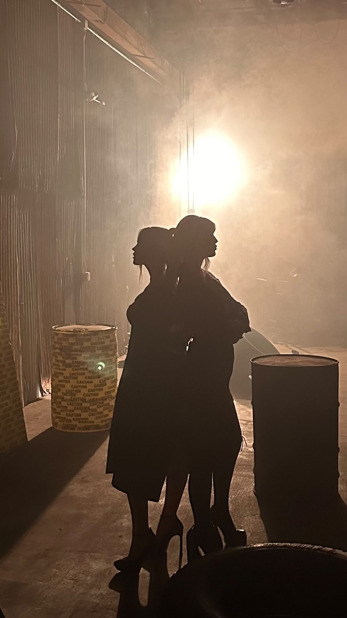 two women stand backtoback in silhouette. both wear dresses and high heels. they are in a smoky warehouse setting with barrels nearby, likely on a stage.