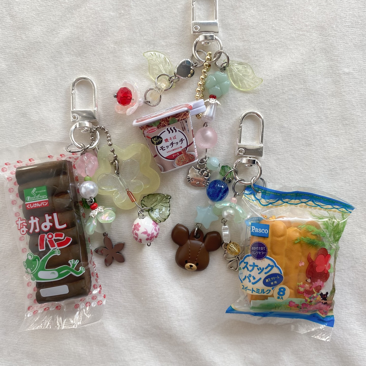 the image shows three keychains with various miniature charms attached to them. the keychains are displayed on a white surface. one keychain features a miniature package of japanese "nakayoshi pan" bread with a cartoon frog on it, along with beads and a small flower charm. another keychain includes a miniature cup of instant noodles, colorful beads, and leafshaped charms. the third keychain has a miniature package of "pasco snack pan" bread with cartoon characters on it, along with beads and a teddy bear charm. the keychains are silver and have clasp closures.