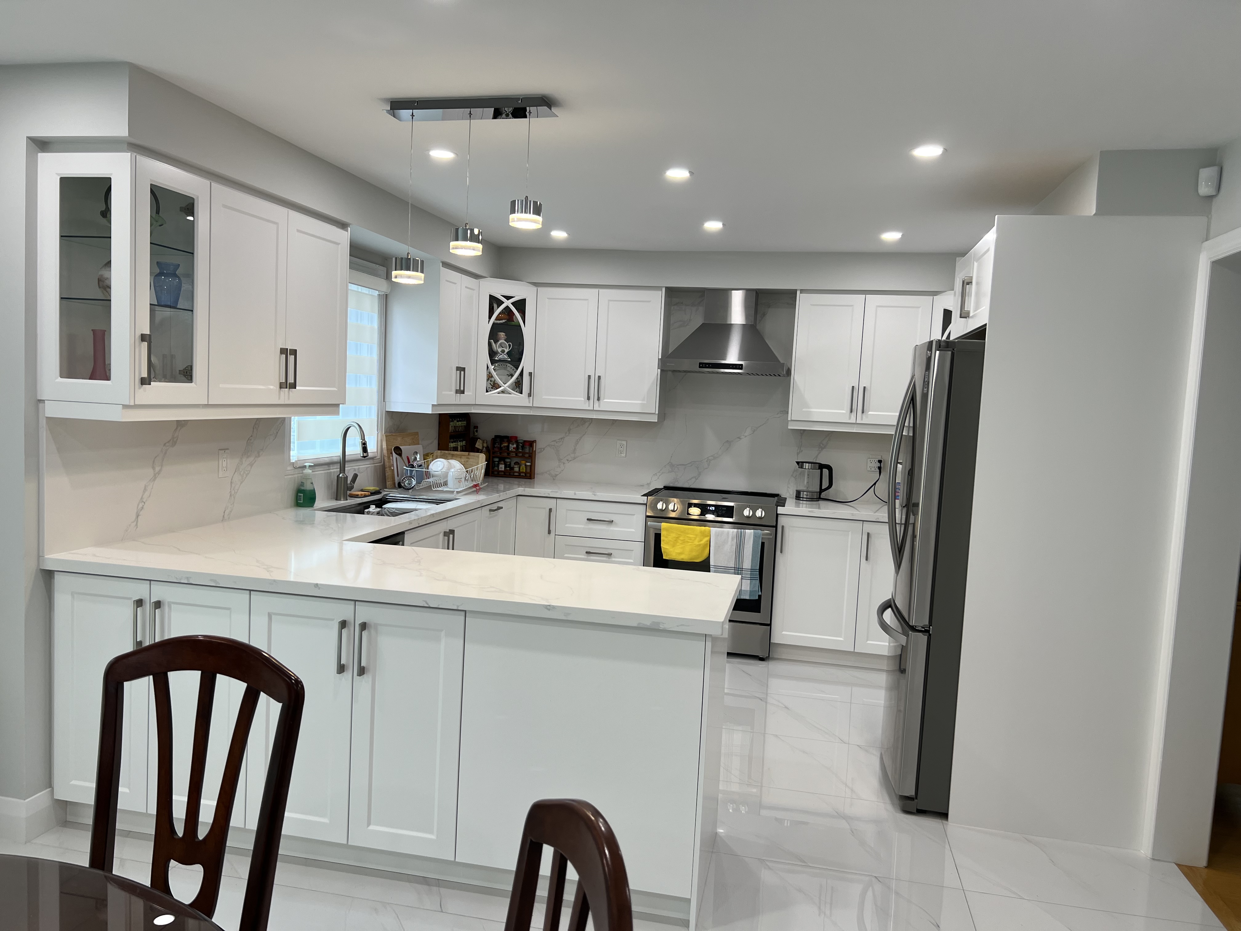 this is a photo of a bright and spacious modern kitchen. the kitchen features white shakerstyle cabinets, quartz countertops, and marble backsplash.  stainless steel appliances, including a refrigerator, oven and range hood, are integrated into the design. the kitchen island provides extra counter space and seating. pendant lights and recessed lighting illuminate the space. the overall style is contemporary and clean.  dark wooden dining chairs are partially visible in the foreground, suggesting a dining area adjacent to the kitchen.  the flooring appears to be large format marble tiles.