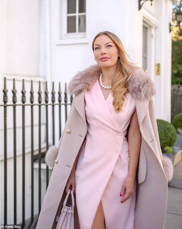a beautiful blonde woman is posing outdoors, wearing a pale pink dress and a light beige coat with a fur collar. she is holding a handbag and looks elegant and classy. the setting is a city street with a white building in the background.
