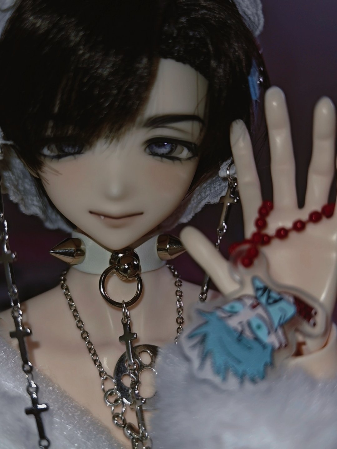 this is a closeup portrait of a doll with long black hair styled in a side part. she's wearing a white choker with silver spikes and a necklace with multiple chains, charms, and a large pendant. she's also wearing bracelets on her wrist and rings on her fingers. her eyes are wide and blue, and her lips are slightly parted. her expression is innocent and sweet. she's wearing a white lace collar and her hair is styled to the side with some strands falling around her face. the background is white.