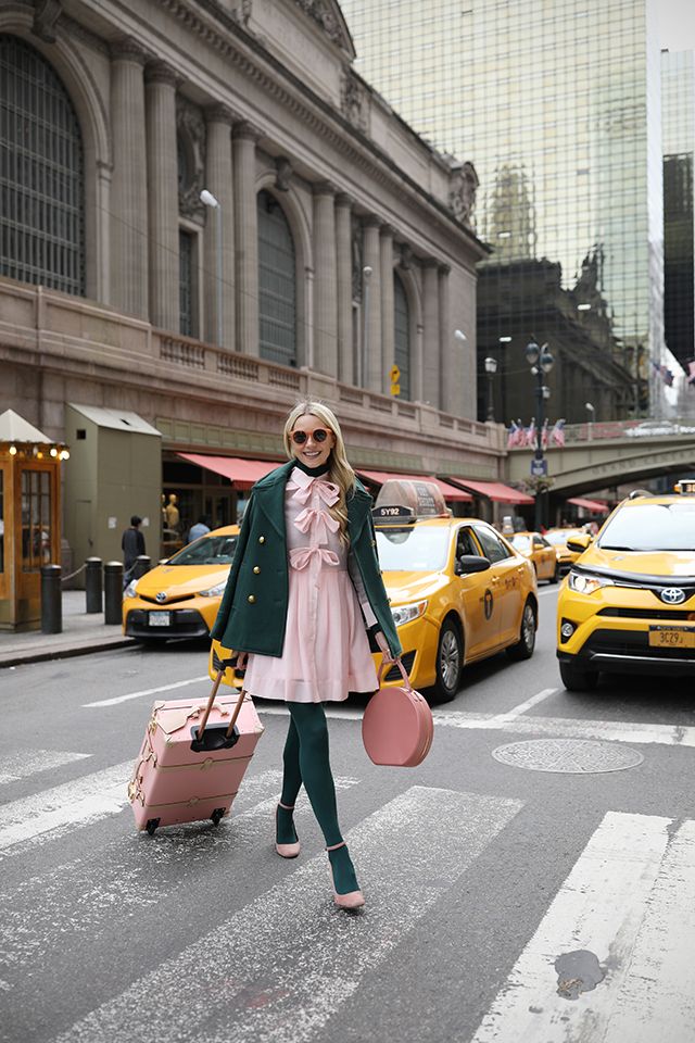 a stylish blonde woman in a green coat and pink dress is crossing a street in new york city, pulling a pink suitcase and carrying a pink handbag.  she's walking past yellow taxis in front of grand central terminal. the overall style is chic and elegant, reflecting an autumnal or spring aesthetic.